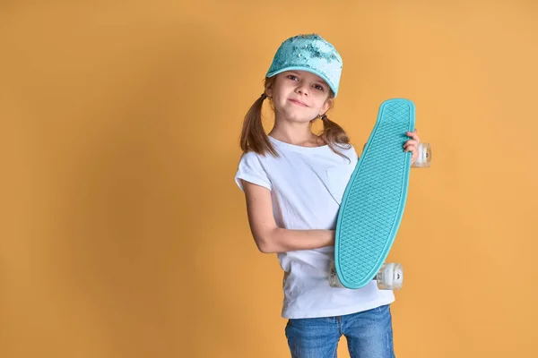 Stylish girl wearing white t-shirt, blue jeans and green cap, holding skateboard over yellow background — Stock Photo, Image