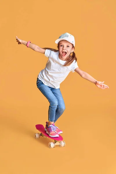 Snygg rolig flicka bär vit t-shirt, blå jeans och sneakers, står på skateboard över gul bakgrund — Stockfoto