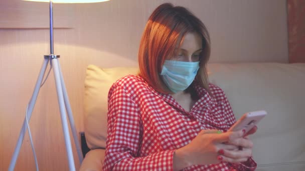 Mujer europea joven con pijama, máscara facial quirúrgica permanecer en casa durante el coronavirus, brote de covid-19 y buscando noticias o aplicación en el teléfono inteligente — Vídeos de Stock