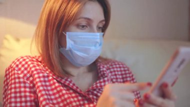 Cerrahi maske takan genç Avrupalı kadın Coronavirus, covid-19 salgını sırasında evde kalıyor ve akıllı telefondan haber veya uygulama izliyor