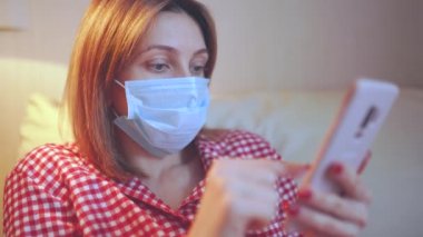 Cerrahi maske takan genç Avrupalı kadın Coronavirus, covid-19 salgını sırasında evde kalıyor ve akıllı telefondan haber veya uygulama izliyor