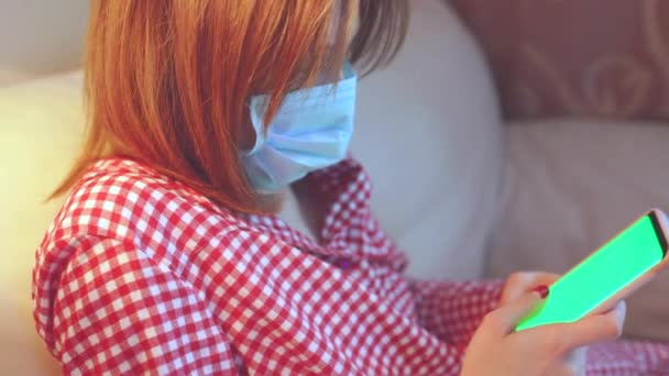 Joven mujer europea con máscara facial quirúrgica permanecer en casa durante el coronavirus, covid-19 brote y buscando noticias o aplicación en el teléfono inteligente — Vídeos de Stock