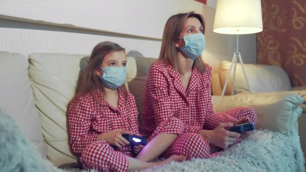 Woman and young girl wearing pajamas and medical protective masks sitting on sofa in living room with video game controllers at home isolation auto quarantine, covid-19 — Stock Video