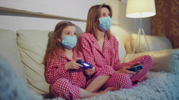 Woman and young girl wearing pajamas and medical protective masks sitting on sofa in living room with video game controllers at home isolation auto quarantine, covid-19 — Stock Video