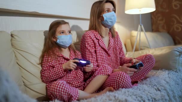 Mujer y niña con pijamas y máscaras protectoras médicas sentadas en el sofá en la sala de estar con controladores de videojuegos en casa de aislamiento de cuarentena automática, covid-19 — Vídeos de Stock