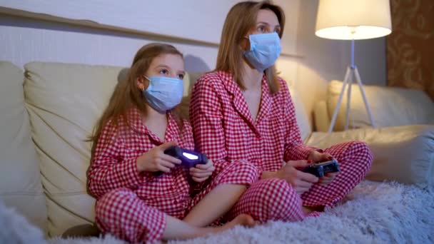 Woman and young girl wearing pajamas and medical protective masks sitting on sofa in living room with video game controllers at home isolation auto quarantine, covid-19 — Stock Video