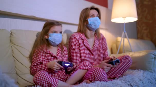 Woman and young girl wearing pajamas and medical protective masks sitting on sofa in living room with video game controllers at home isolation auto quarantine, covid-19 — Stock Video