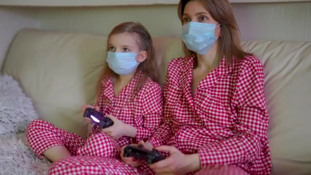 Woman and young girl wearing pajamas and medical protective masks sitting on sofa in living room with video game controllers at home isolation auto quarantine, covid-19 — Stock Video