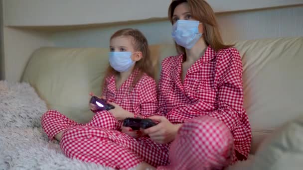 Woman and young girl wearing pajamas and medical protective masks sitting on sofa in living room with video game controllers at home isolation auto quarantine, covid-19 — Stock Video