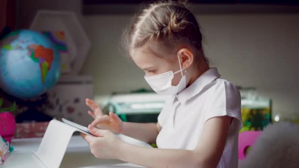 Ragazza preadolescente caucasica con maschera medica sul viso concentrata sul suo compito con tablet. Concetto di apprendimento a distanza in isolamento mentre coronavirus — Video Stock