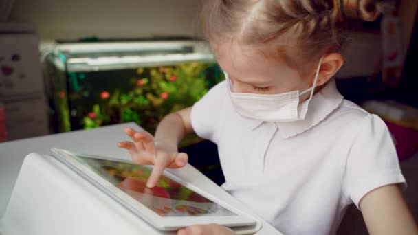 Ragazza preadolescente caucasica con maschera medica sul viso concentrata sul suo compito con tablet. Concetto di apprendimento a distanza in isolamento mentre coronavirus — Video Stock