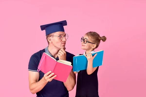 Άντρας σε ακαδημαϊκό καπέλο κρατώντας βιβλίο, μελέτη μαζί με χαριτωμένο preteen κορίτσι σε σχολική στολή. Πατέρας και κόρη απομονωμένοι σε ροζ φόντο στο στούντιο. Η έννοια της οικογενειακής ημέρας πατρότητας — Φωτογραφία Αρχείου