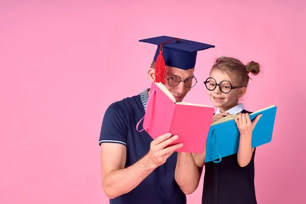 Homem em chapéu acadêmico segurando livro, estudar em conjunto com a menina pré-adolescente bonito em uniforme escolar. Pai, filha isolada em fundo rosa no estúdio. Amor dia da família paternidade conceito de infância — Fotografia de Stock