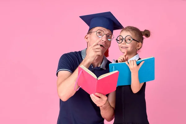 Man in academic hat holding book, study together with cute preteen girl in school uniform. Father, daughter isolated on pink background in studio. Love family day parenthood childhood concept — Stock Photo, Image