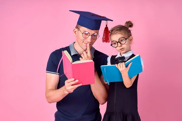 学術的な帽子の男は本を保持し、学校の制服でかわいい十代の女の子と一緒に勉強します。娘さんはスタジオでピンクの背景に隔離されていました。家族の日の親の子供時代のコンセプト — ストック写真