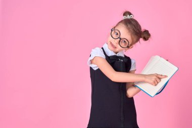 Üniformalı bir ilkokul öğrencisi, gözlüksüz yuvarlak gözlüklü elinde bir not defteri tutuyor ve stüdyoda pembe bir arka plandaki boş bir sayfayı işaret ediyor.