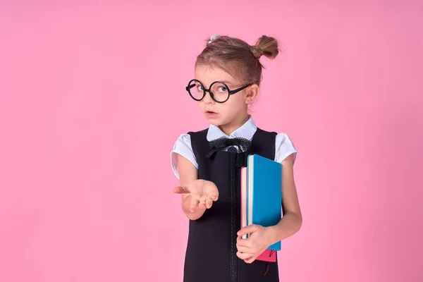 Κορίτσι του δημοτικού σχολείου με στολή, στρογγυλά γυαλιά χωρίς φακούς κρατά ένα σημειωματάριο στα χέρια της και σηκώνει το χέρι της στο πλάι με το συναίσθημα μιας ερώτησης στο πρόσωπό της, θέτοντας σε ροζ φόντο — Φωτογραφία Αρχείου