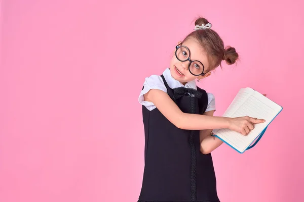 Niña de la escuela primaria en uniforme, gafas redondas sin lentes sostiene un cuaderno en sus manos y señala con el dedo una página en blanco sobre un fondo rosa en el estudio — Foto de Stock