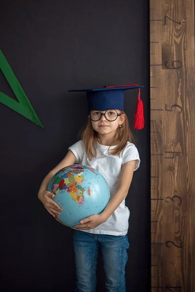 Menina em chapéu acadêmico e óculos arredondados fica na parede preta segurando globo atrás de uma régua, de volta à escola, conceito pré-escolar, criança está se preparando para a escola — Fotografia de Stock