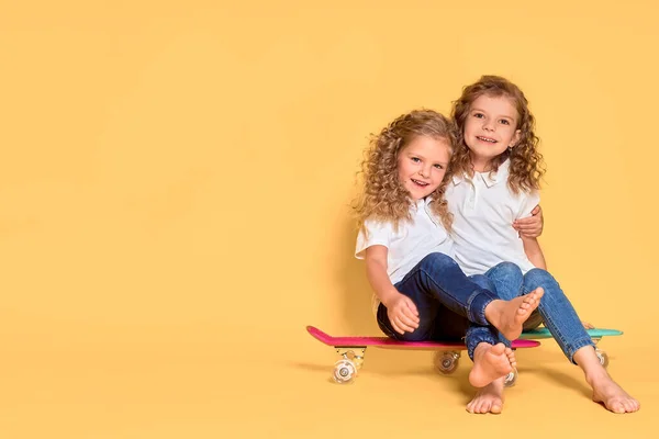 Deux filles actives et heureuses aux cheveux bouclés s'amusent avec un penny board, un visage souriant et une planche à roulettes. Penny board skateboard mignon pour les filles. Allons-y. Fille avec planche de penny fond jaune — Photo