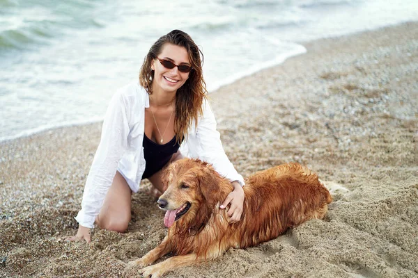 Портрет счастливой молодой женщины, сидящей и обнимающей свою собаку на пляже — стоковое фото