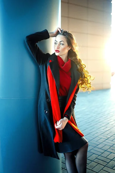 Joven chica hermosa con maquillaje perfecto, labios rojos, con un abrigo oscuro, camisa roja, posando cerca del centro comercial. Acaba de salir de una tienda de ropa de alta gama. — Foto de Stock
