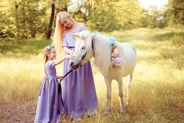 Mãe e filha em vestidos de lavanda semelhantes estão acariciando um cavalo de unicórnio. Prado de verão — Fotografia de Stock