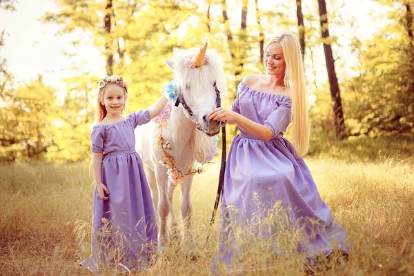 Mãe e filha em vestidos de lavanda semelhantes estão acariciando um cavalo de unicórnio. Prado de verão — Fotografia de Stock