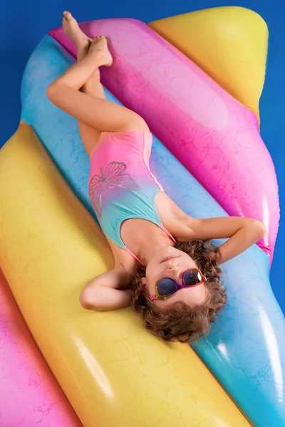 Pretty smiling girl wearing pink and blue swimwear and sunglasses lies, relax on rainbow inflatable mattress. — Stock Photo, Image