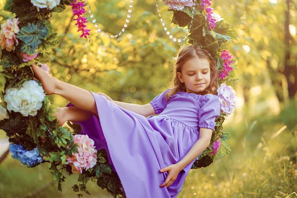 Menina bonito vestindo vestido lilás posando enquanto sentado no anel decorado com flores no verão parque — Fotografia de Stock