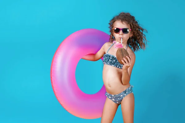 Portret szczęśliwej dziewczynki w stroju kąpielowym w okularach przeciwsłonecznych z nadmuchiwanym pierścieniem i kokosem ze słomką odizolowaną od niebieskiego tła — Zdjęcie stockowe