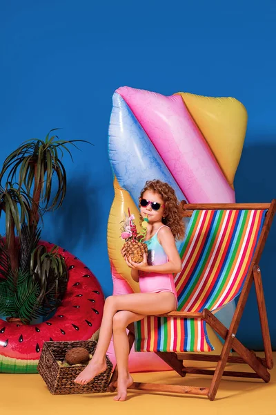 Uśmiechnięta dziewczyna w stroju kąpielowym i okularach przeciwsłonecznych siedząca na tęczowym leżaku trzymająca koktajl ananasowy z kolorowymi słomkami i pokazująca kciuk w górę i opalanie — Zdjęcie stockowe
