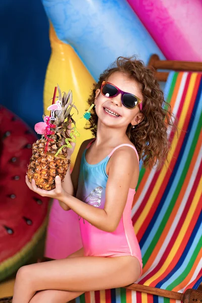 Усміхнена дівчина в купальнику і сонцезахисних окулярах, сидячи в кріслі на веселковій палубі, тримає ананасовий коктейль з різнокольоровими соломинками і показує великий палець вгору і засмагає — стокове фото
