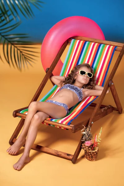 Girl in swimsuit and sunglasses lying in rainbow deck chair with crossed legs and sunbathing — Stock Photo, Image
