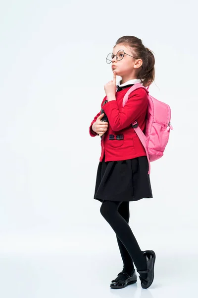 Adorable niña en chaqueta de escuela roja, vestido negro, mochila y gafas redondeadas abraza un libro firmemente y sonríe mientras disfruta y posa sobre fondo de estudio blanco. Aislar — Foto de Stock