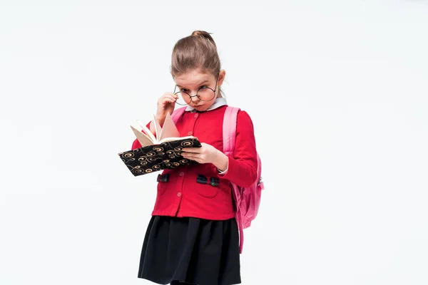 Menina adorável na jaqueta da escola vermelha, vestido preto, mochila e óculos arredondados surpreendentes ou espantados olhando para o livro enquanto posando no fundo do estúdio branco. Isolar — Fotografia de Stock
