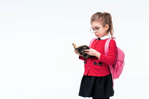 Adorable niña en chaqueta de escuela roja, vestido negro, mochila y gafas redondeadas mirando emocionalmente el libro mientras posa sobre fondo blanco del estudio. Aislar — Foto de Stock