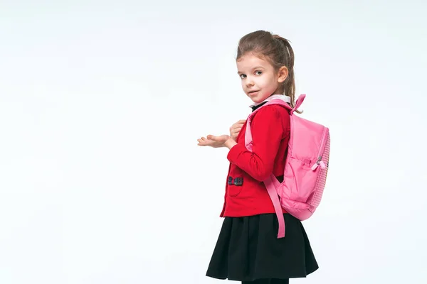 Adorable niña en chaqueta de escuela roja, vestido negro, en las correas de una mochila y sonriendo y mirando a la cámara, posando sobre fondo blanco estudio. Aislar — Foto de Stock