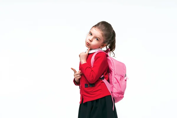 Adorable niña en chaqueta de escuela roja, vestido negro, mochila apuntando al espacio vacío mientras posa sobre fondo blanco del estudio. Aislar —  Fotos de Stock