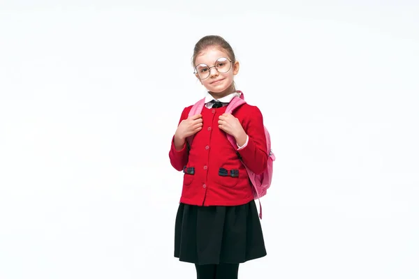 Чарівна маленька дівчинка в червоній шкільній куртці, чорна сукня, округлі окуляри тримаються на ремінцях рюкзака, посміхаючись і дивлячись на камеру, позує на білому фоні студії. ізольовані — стокове фото