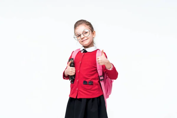 Menina adorável em casaco vermelho escola, vestido preto, óculos arredondados e mochila mostrando polegar para cima enquanto posando no fundo do estúdio branco. Isolar — Fotografia de Stock