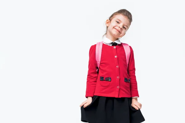 Menina adorável na jaqueta da escola vermelha, vestido preto, mochila timidamente se apega a uma saia e sorrir enquanto posando no fundo do estúdio branco. Isolar — Fotografia de Stock