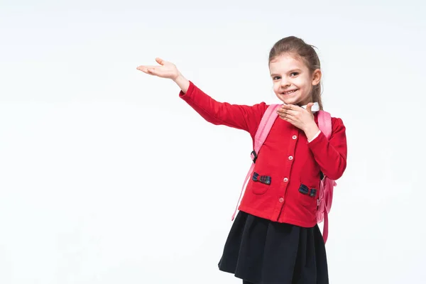 Чарівна маленька дівчинка в червоній шкільній куртці, чорна сукня, рюкзак, що вказує на міміку простору, позуючи на білому фоні студії. ізольовані — стокове фото
