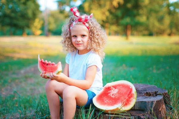 Šťastné kudrnaté dítě dívka sedí na pařezu a jíst vodní meloun venku v jarním parku proti přirozenému slunné rozmazané pozadí — Stock fotografie