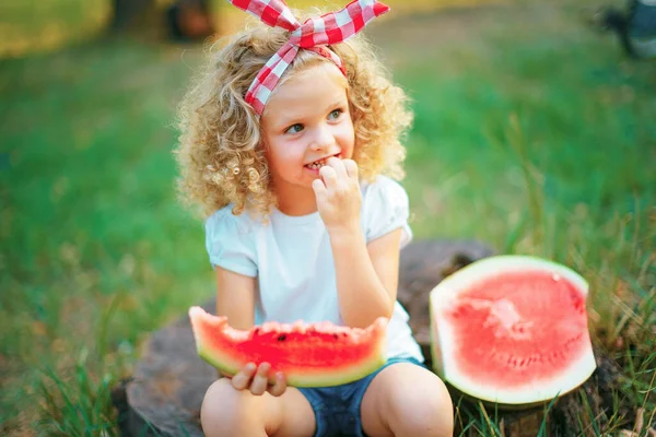 Mutlu kıvırcık kız çocuğu kütüğün üzerinde oturuyor ve bahar parkında karpuz yiyor. — Stok fotoğraf