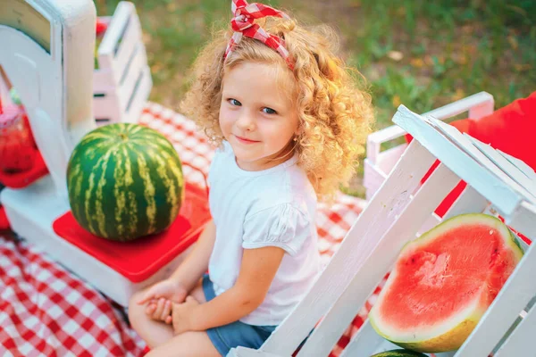 Šťastné kudrnaté dítě dívka sedí na pařezu a jíst vodní meloun venku v jarním parku proti přirozenému slunné rozmazané pozadí — Stock fotografie