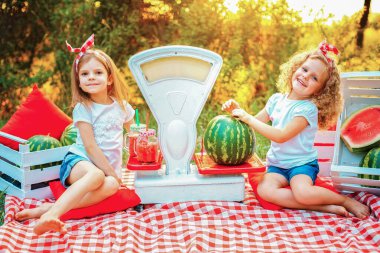 Beyaz tişörtlü ve kırmızı bantlı iki küçük kız doğada bir halıya oturmuş ellerinde meyve suyu dolu bir karpuz ve kavanozla eski moda bir terazinin üzerinde duruyorlar. Satış konsepti