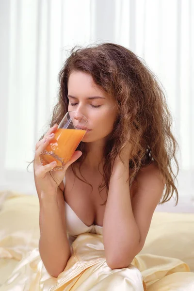 Mujer alegre bebiendo un jugo de naranja sentada en su cama en casa —  Fotos de Stock