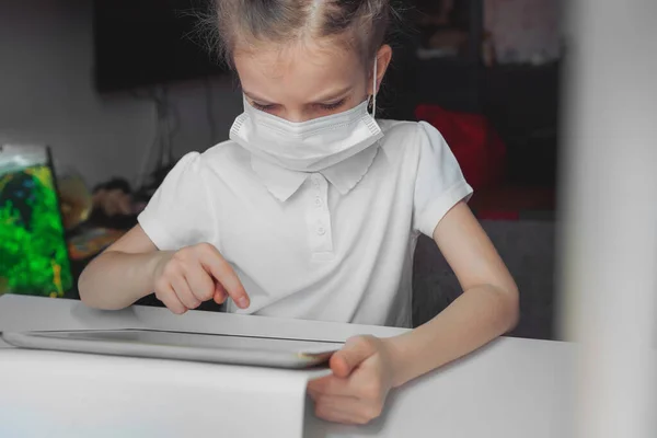 Kaukasiska preteen flicka med medicinsk mask på ansiktet koncentrerade sig på sin uppgift med tablett. Begreppet distansundervisning isolerat medan coronavirus — Stockfoto