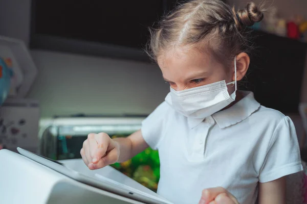 Kaukasiska preteen flicka med medicinsk mask på ansiktet koncentrerade sig på sin uppgift med tablett. Begreppet distansundervisning isolerat medan coronavirus — Stockfoto